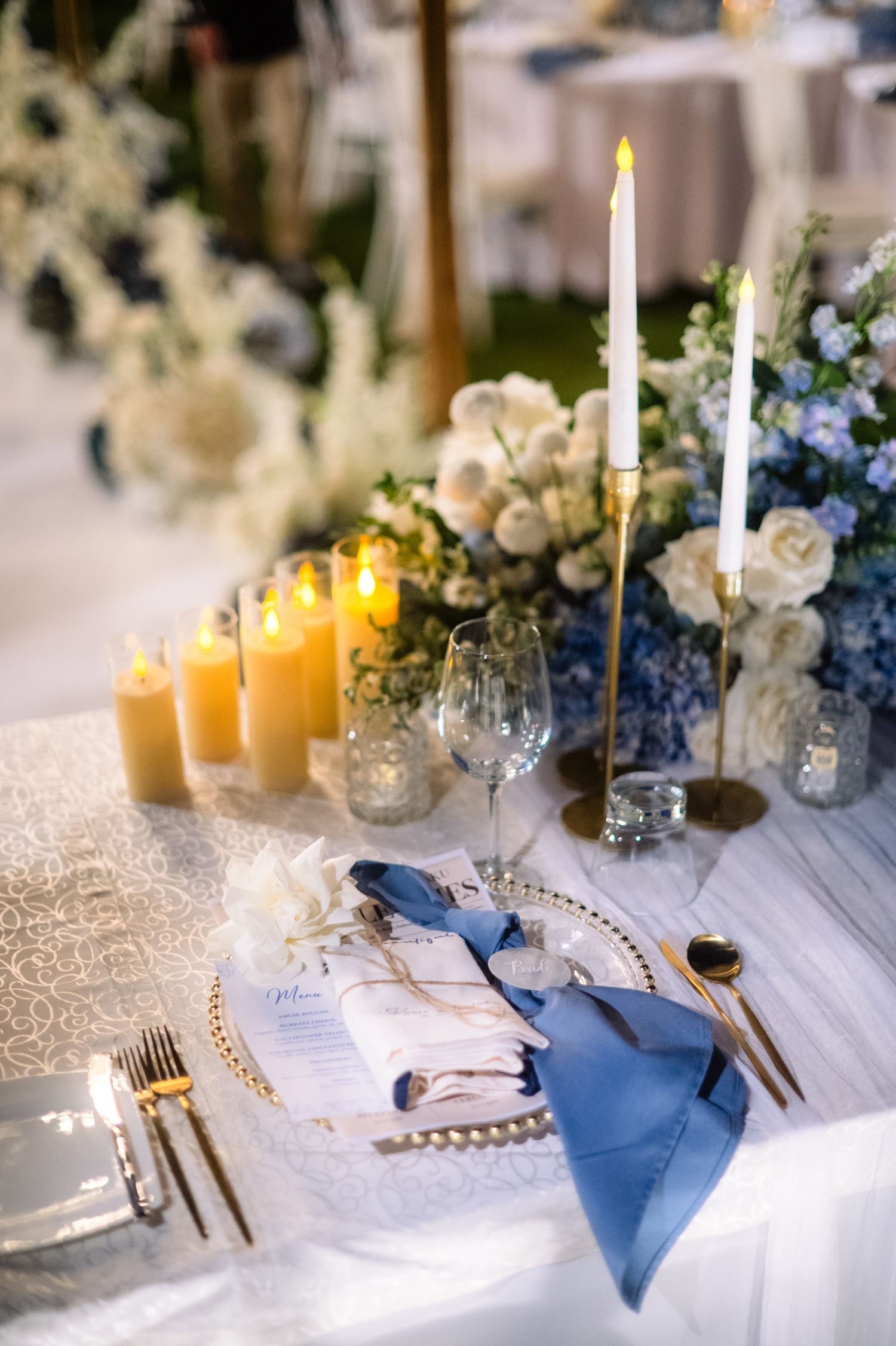 Elegant wedding table setting with blue napkin, gold flatware, taper candles, pillar candles, and white and blue floral centerpieces.