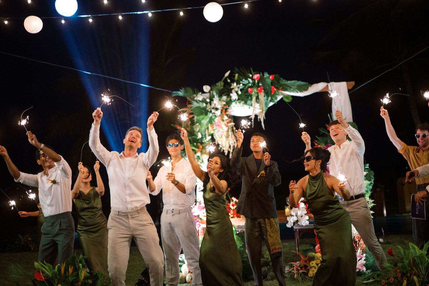 Wedding guests celebrate joyfully with sparklers at an outdoor evening tropical wedding reception.