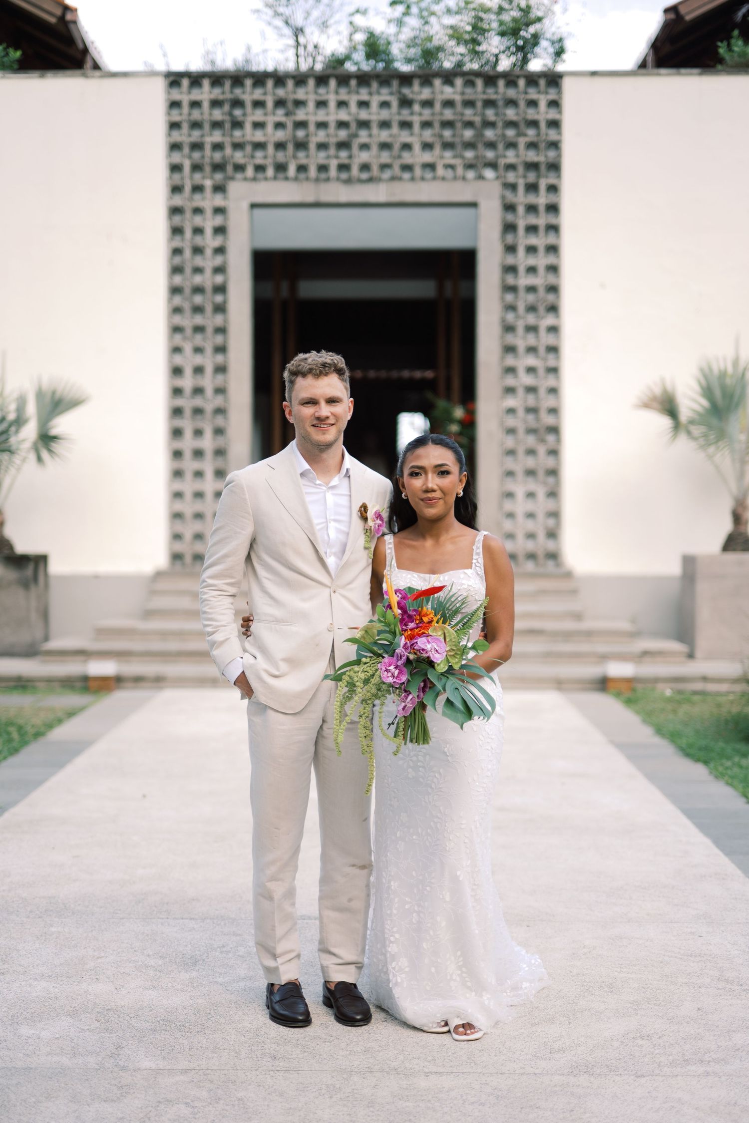 Bride in white gown holding tropical bouquet stands with groom in beige suit at elegant outdoor wedding venue.
