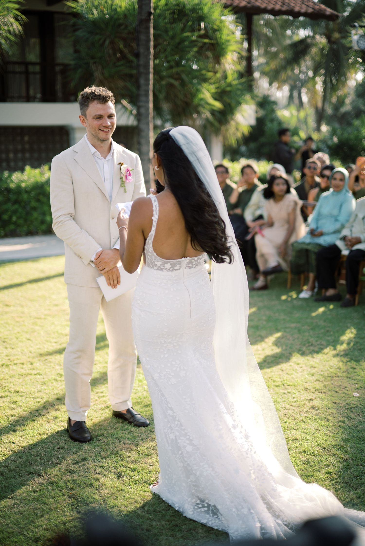 Groom in cream suit smiles at bride during outdoor wedding ceremony with guests seated.