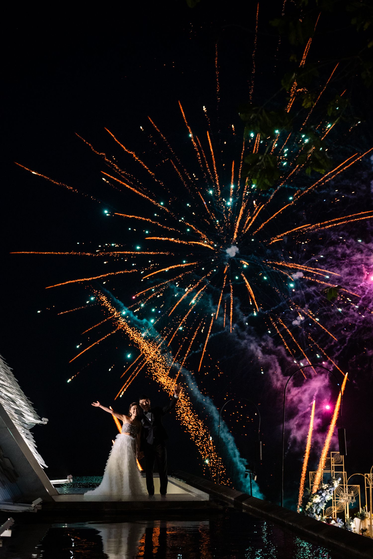 Bride in a white gown and groom in a dark suit stand beside a reflective pool as orange and purple fireworks light up the night sky.