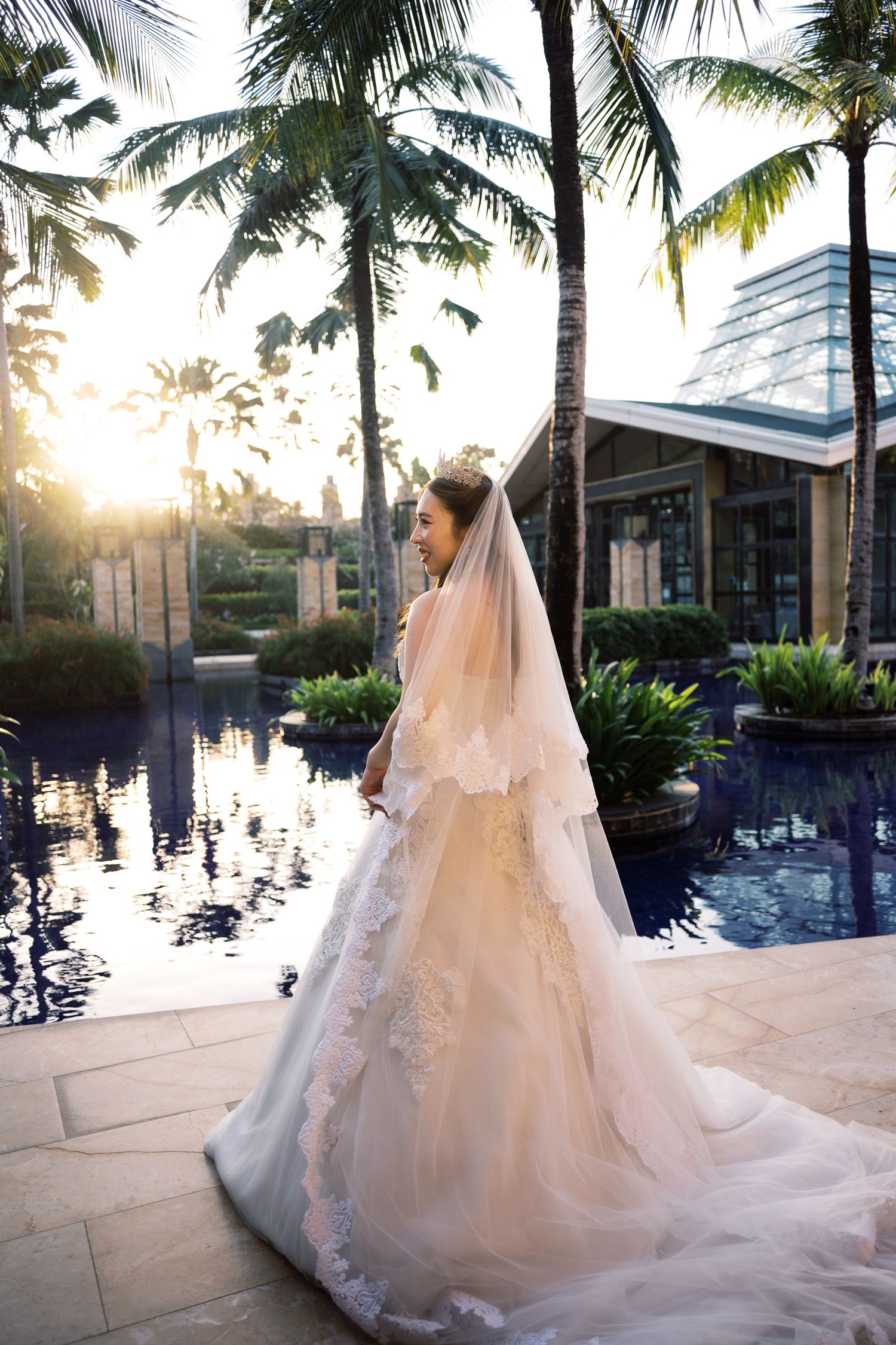 Bride in lace wedding gown and veil poses by tropical pool at sunset with palm trees.