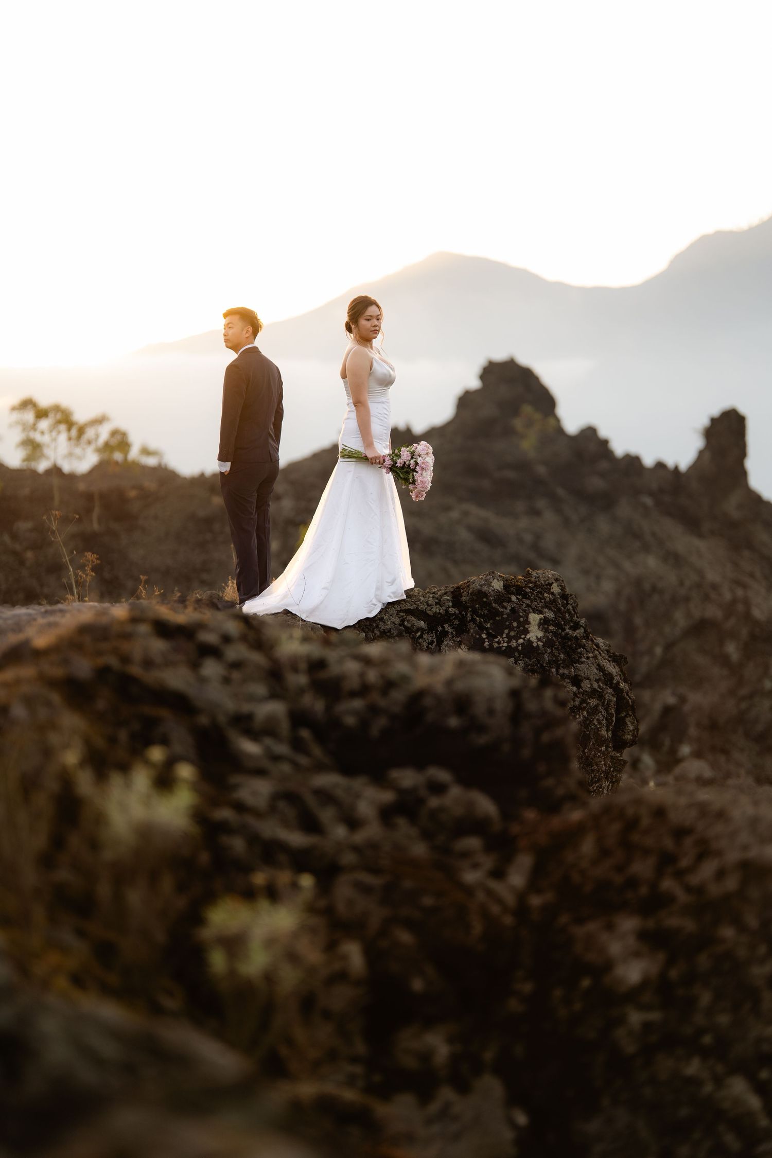 Bride and groom standing on volcanic rocks at sunset with mountain backdrop on their wedding day.