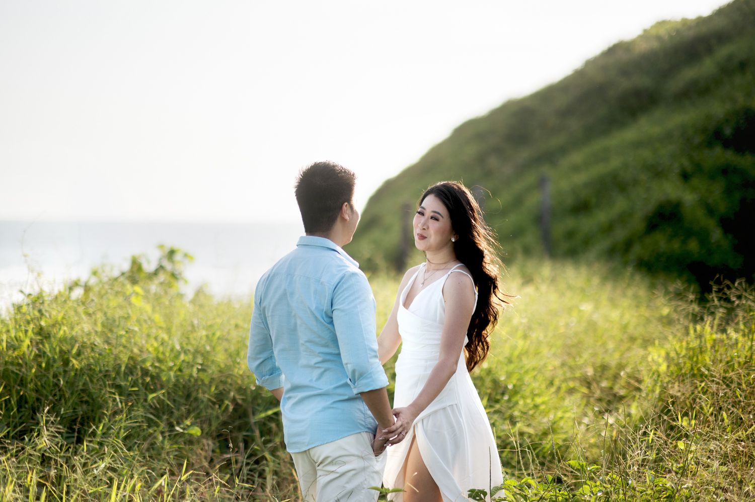 Couple holding hands in a lush green meadow near the ocean, smiling at each other in golden hour light.