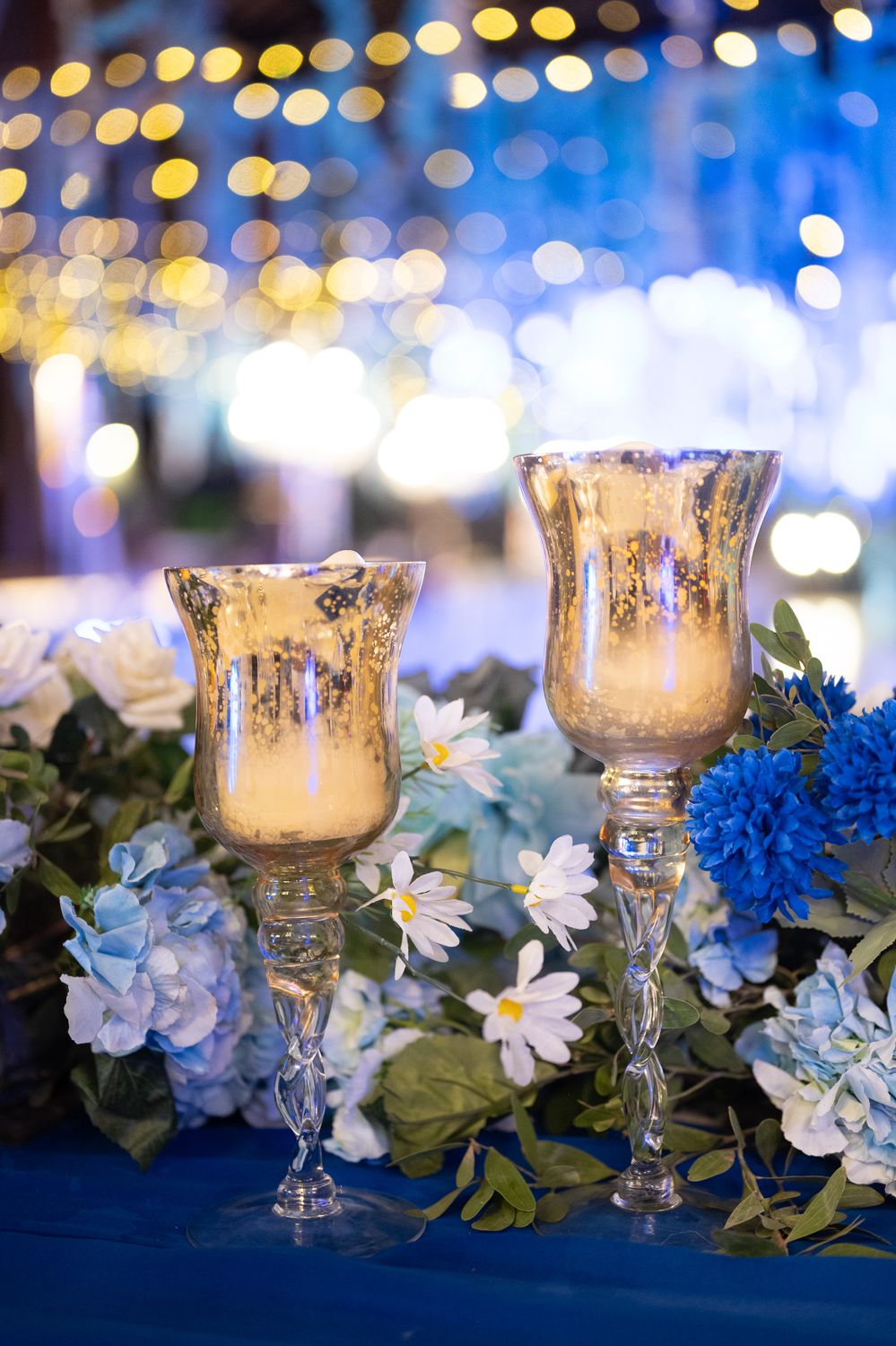 Two gold mercury glass candle holders surrounded by blue hydrangeas and white daisies on a blue tablecloth with bokeh lights.