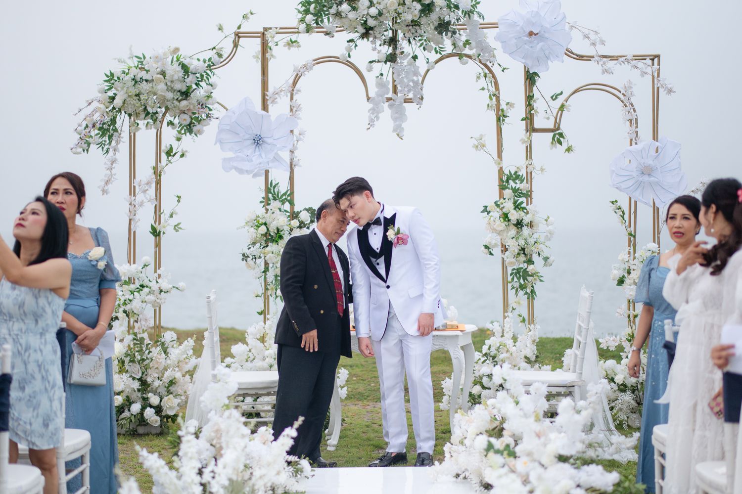 Couple shares a kiss at an elegant outdoor wedding ceremony with white floral arch and guests in formal attire.
