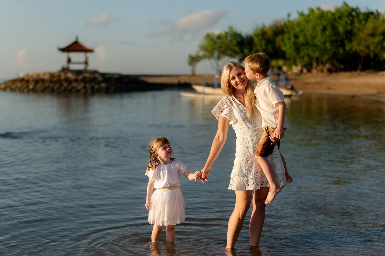 Blonde mother holding son while daughter stands in shallow water on a tropical beach at sunset.