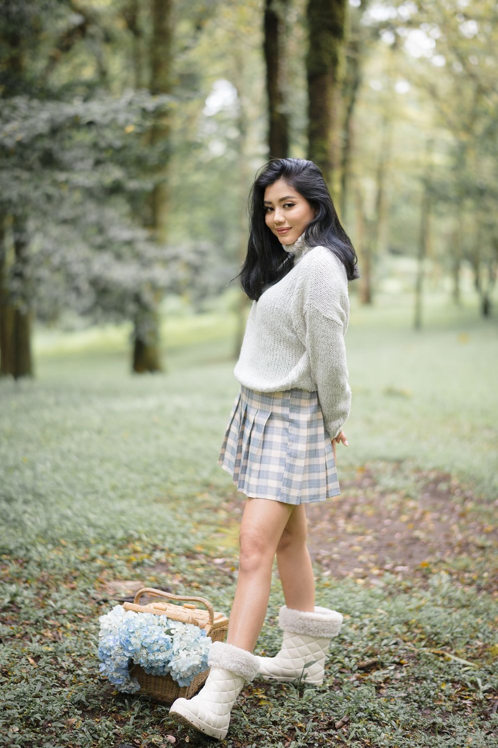 Young Asian woman in plaid skirt and knit sweater poses in a lush green forest with a flower basket nearby.