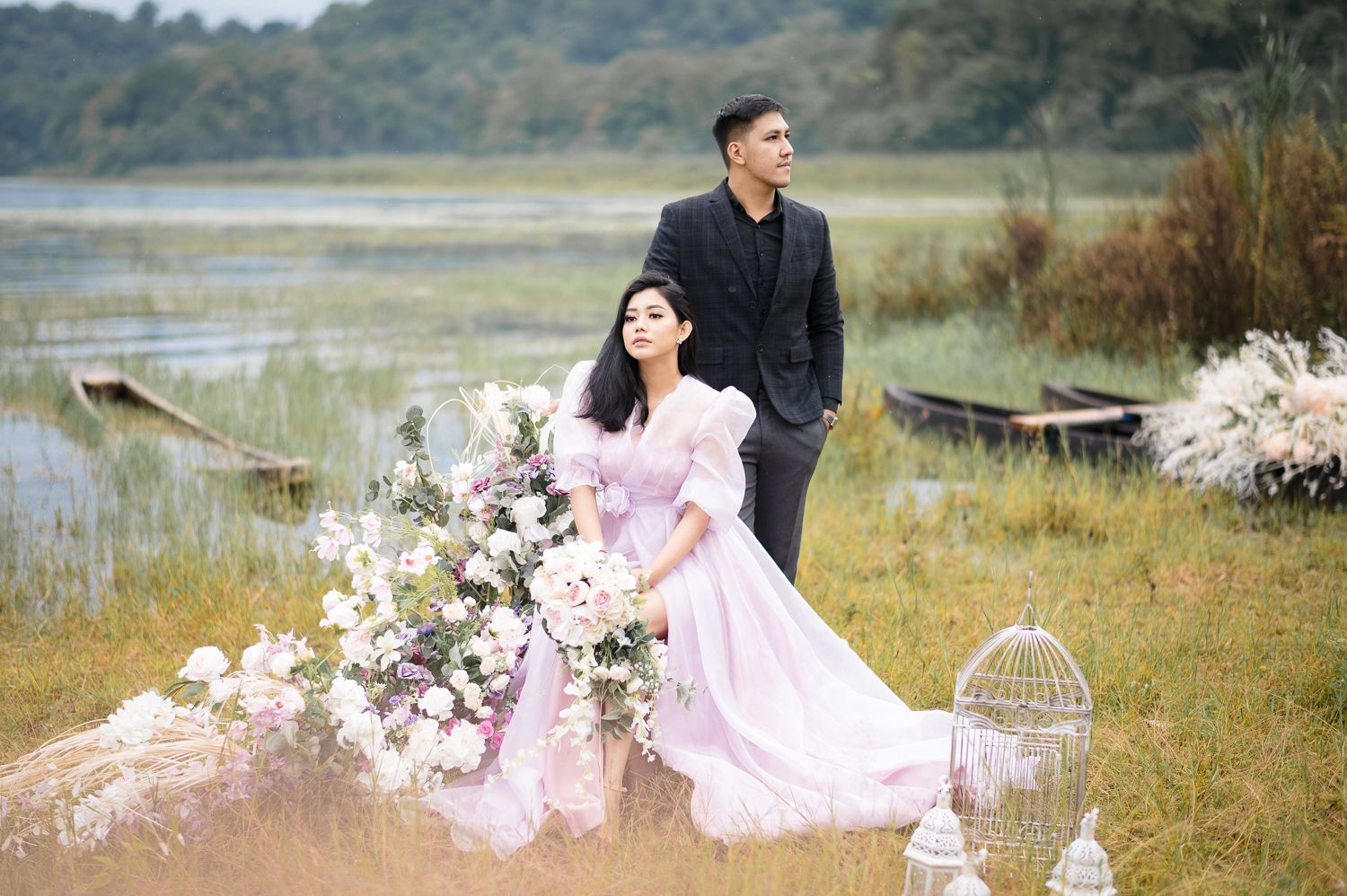 Couple in elegant attire posing near a lakeside with floral arrangements and a birdcage in a romantic outdoor setting.