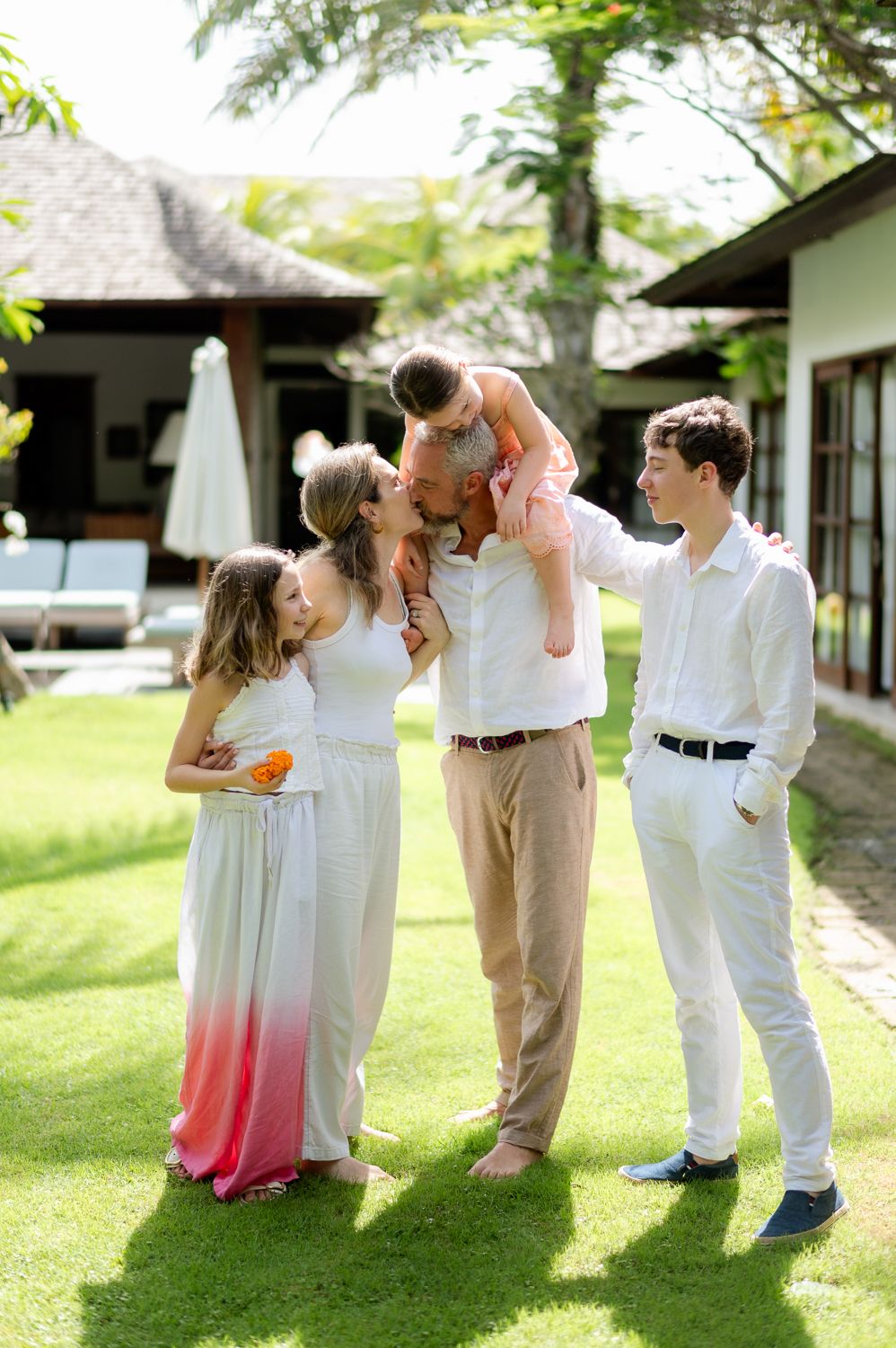Family of five sharing a joyful moment on green lawn outside a tropical villa on a sunny day.
