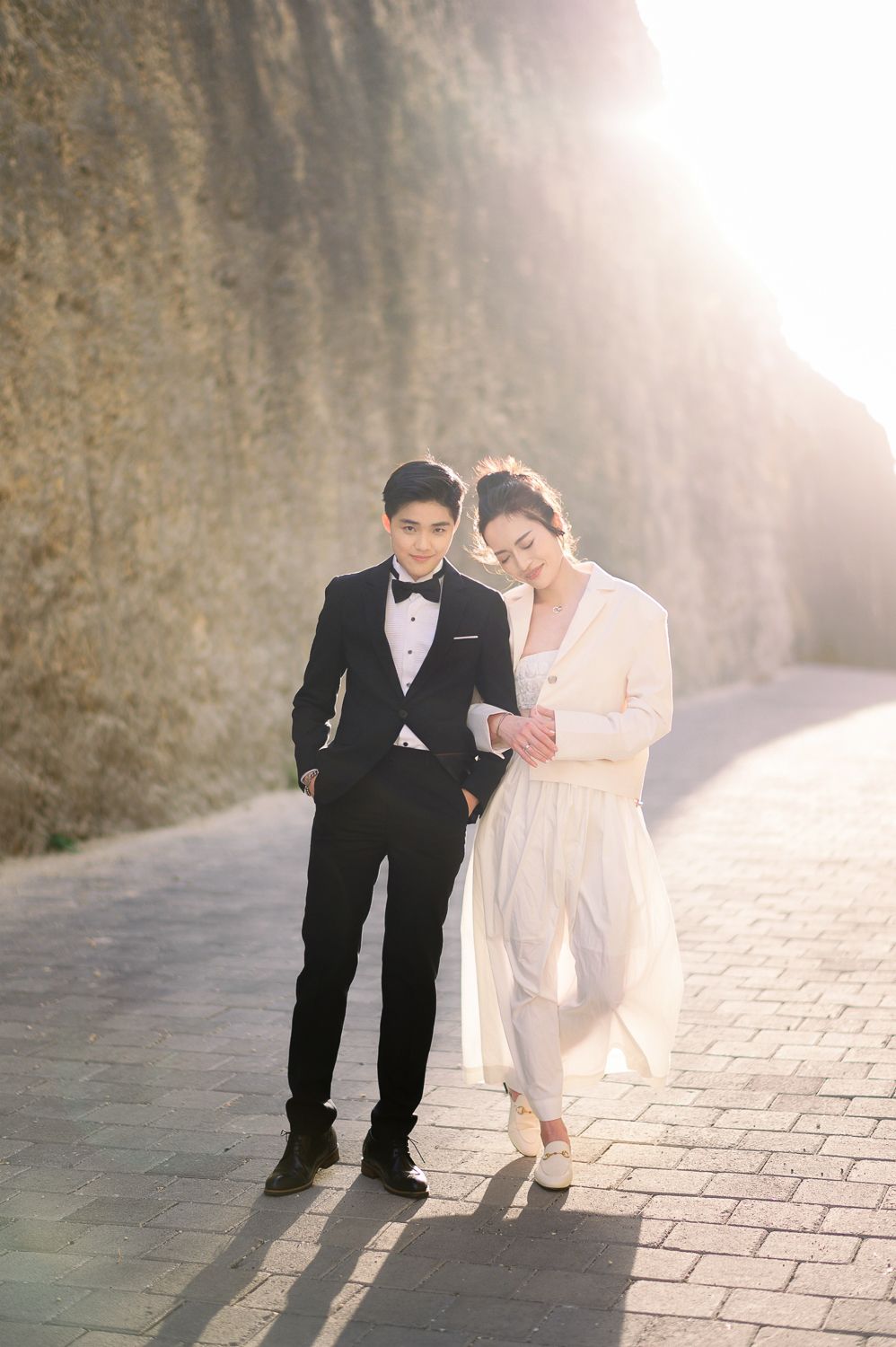 Couple in formal wedding attire posing on a sunlit stone pathway beside a rocky cliff wall.