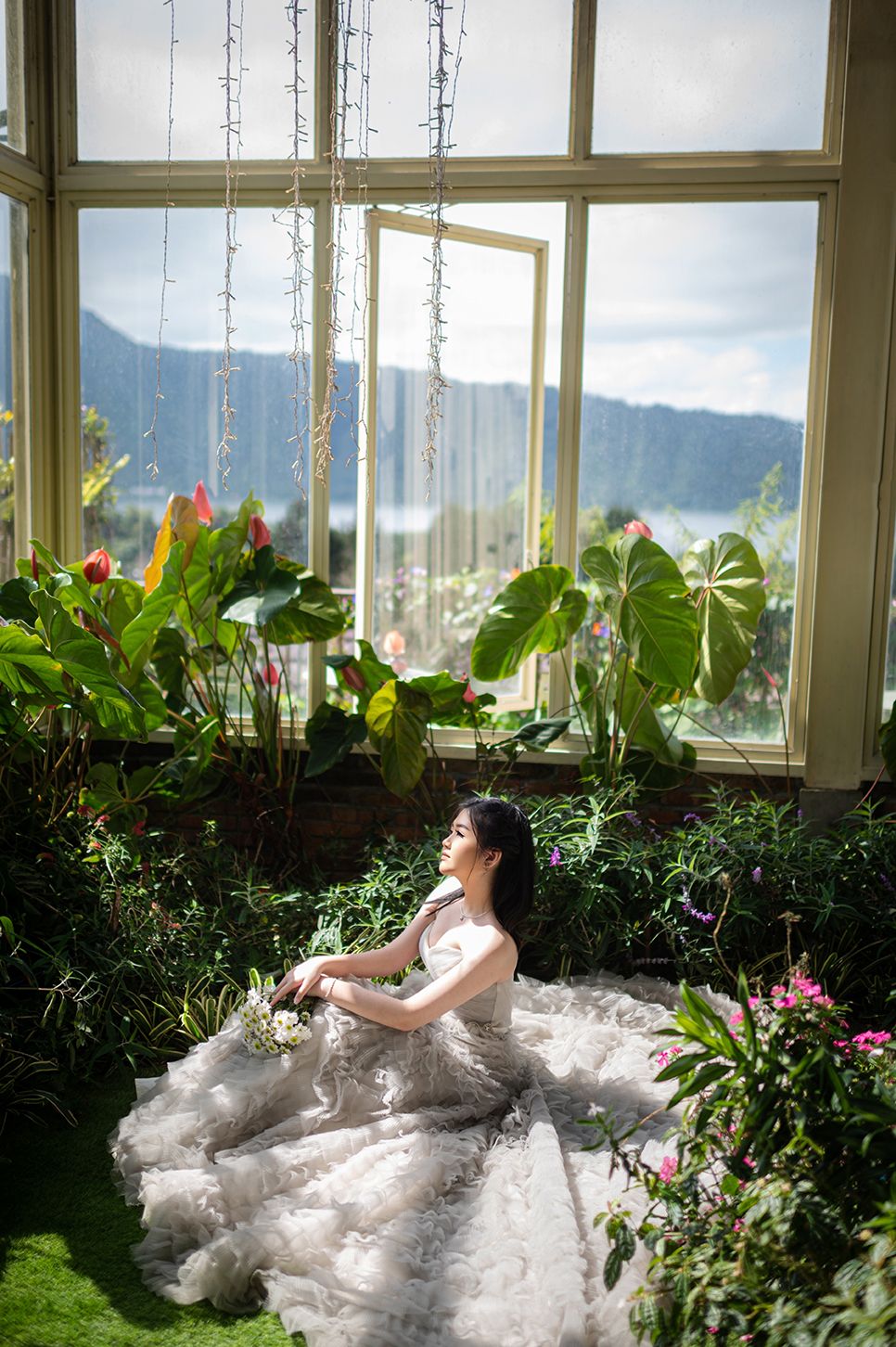 Bride in a flowing gown sits among lush tropical plants in a sunlit greenhouse with mountain views.