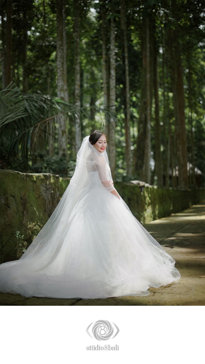 Bride in a flowing white ball gown with veil poses on a stone path surrounded by tall trees in Bali.