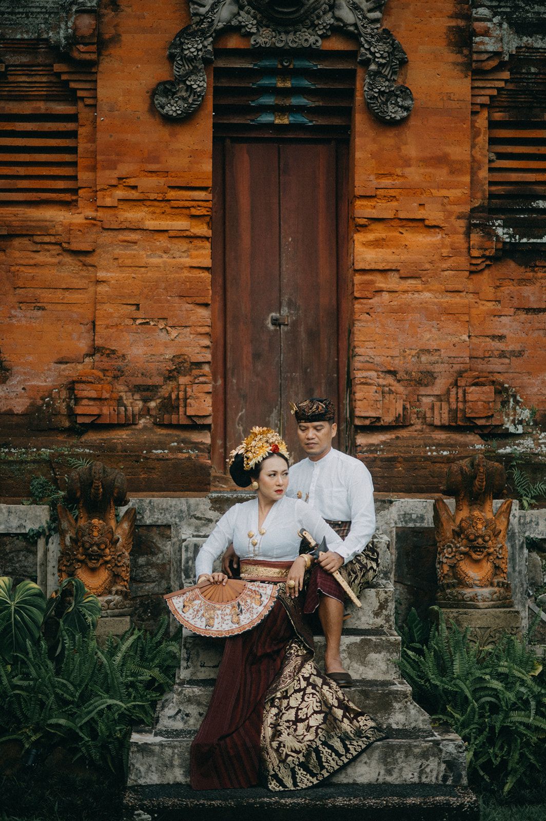 Balinese couple in traditional attire posing before an ornate temple door with stone carvings and lush greenery.