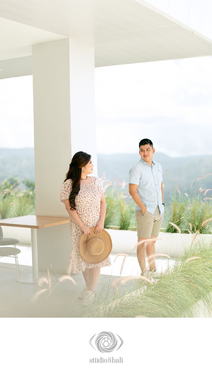 A couple poses on a modern white terrace surrounded by lush greenery and hills, woman in floral dress holding a straw hat.