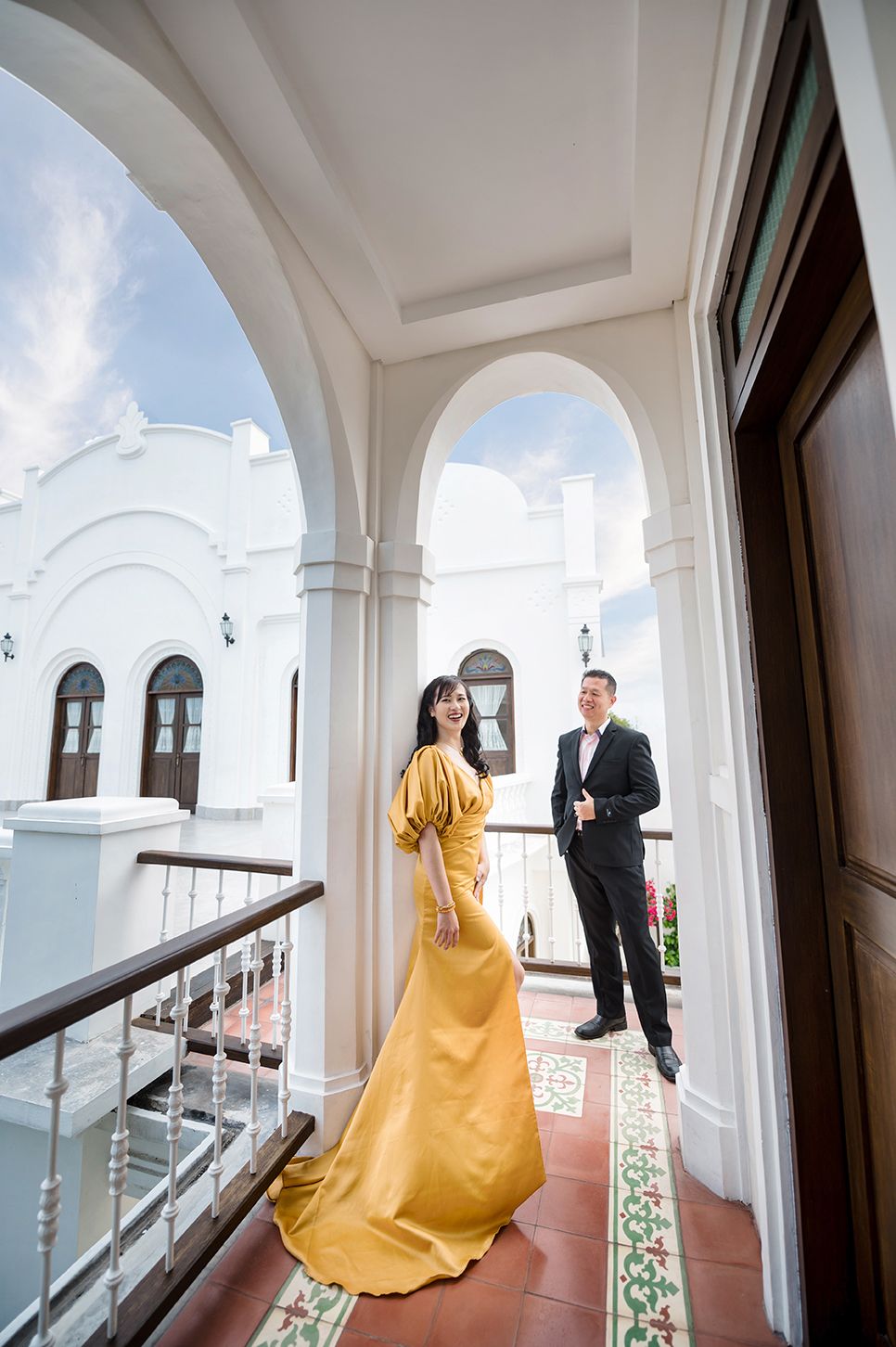 Couple posing on elegant white colonial balcony, woman in gold gown, man in black suit.