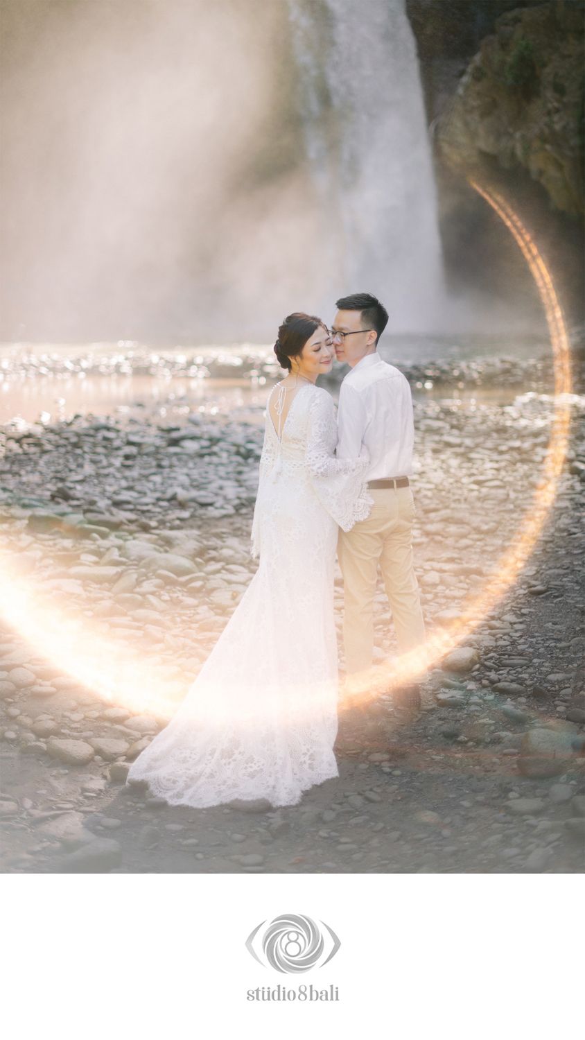 Couple in white attire embracing near a waterfall with a glowing circular light effect, captured by Studio 8 Bali.