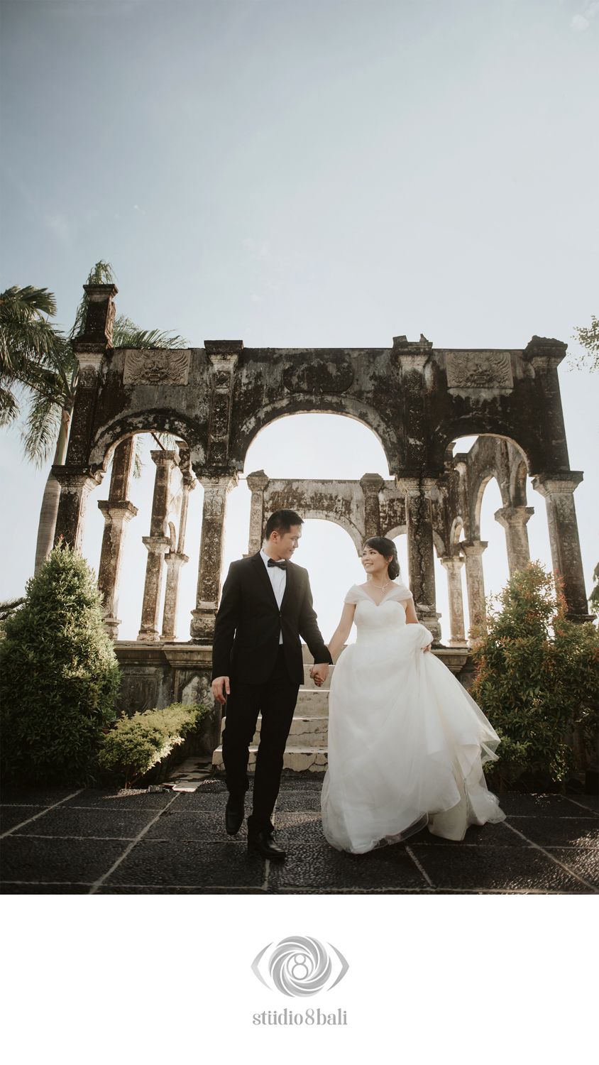 Couple in wedding attire holding hands before ancient stone arch ruins in Bali, Indonesia.