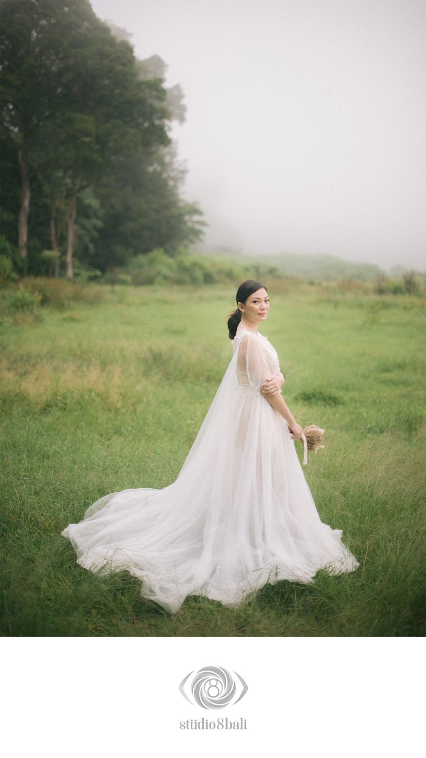 Bride in flowing white gown with long veil stands in misty green field holding bouquet, captured by Studio 8 Bali.