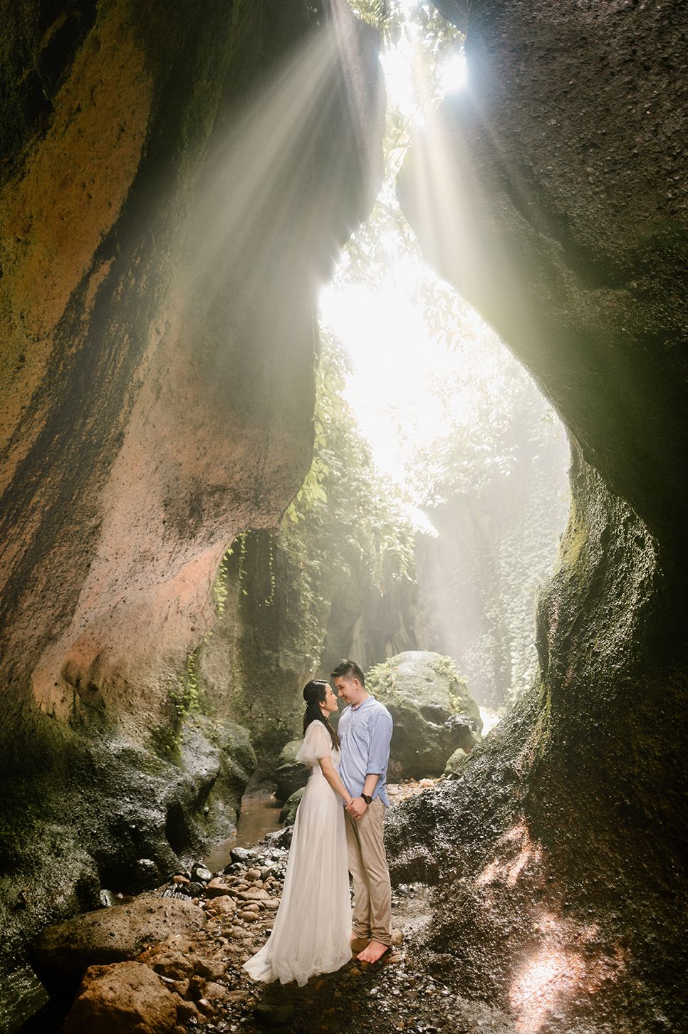 Couple embracing in a sunlit cave with dramatic light rays, surrounded by rocky walls and lush greenery.