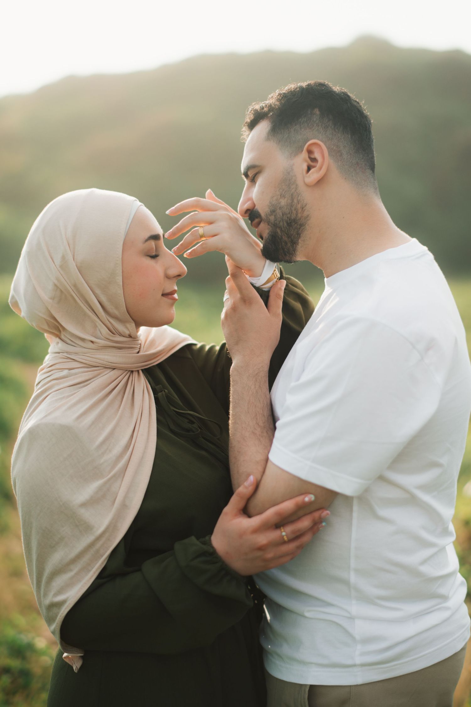 A couple sharing a tender moment outdoors, woman in hijab gently touching man's face in golden sunlight.