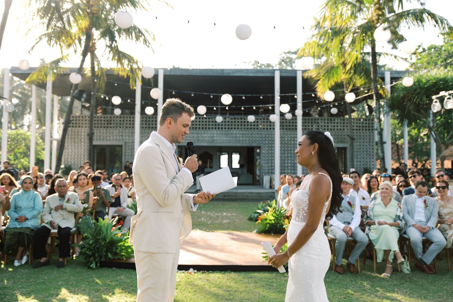 Groom reads vows to bride before large seated wedding crowd at outdoor tropical ceremony.