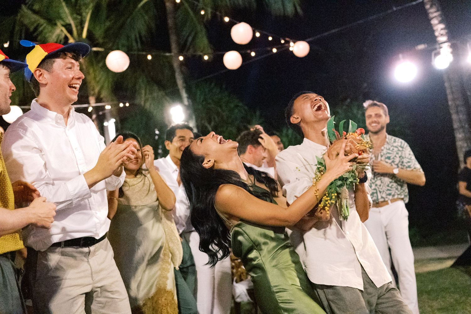 Excited wedding guests laugh and celebrate bouquet toss at outdoor tropical reception.