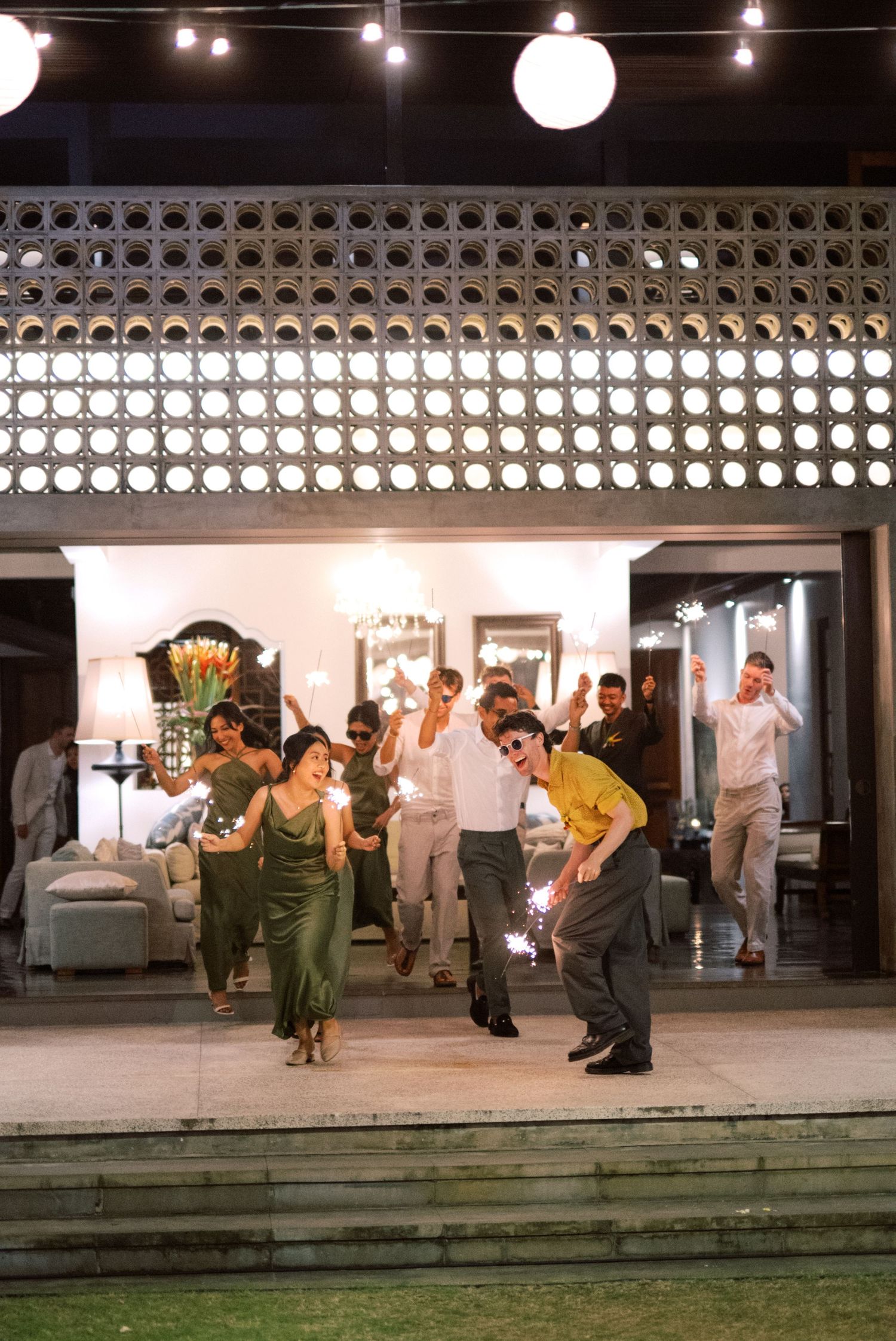 Wedding guests celebrate with sparklers outside decorated venue at night during lively outdoor reception party.