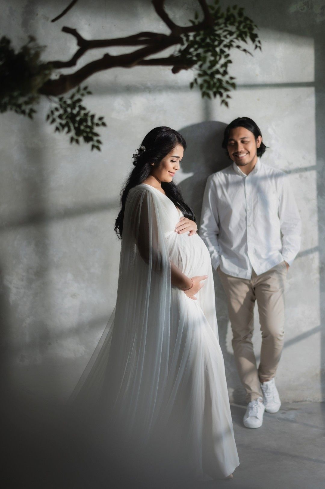 Pregnant woman in white maternity gown poses with smiling man in white shirt for elegant maternity photo shoot.