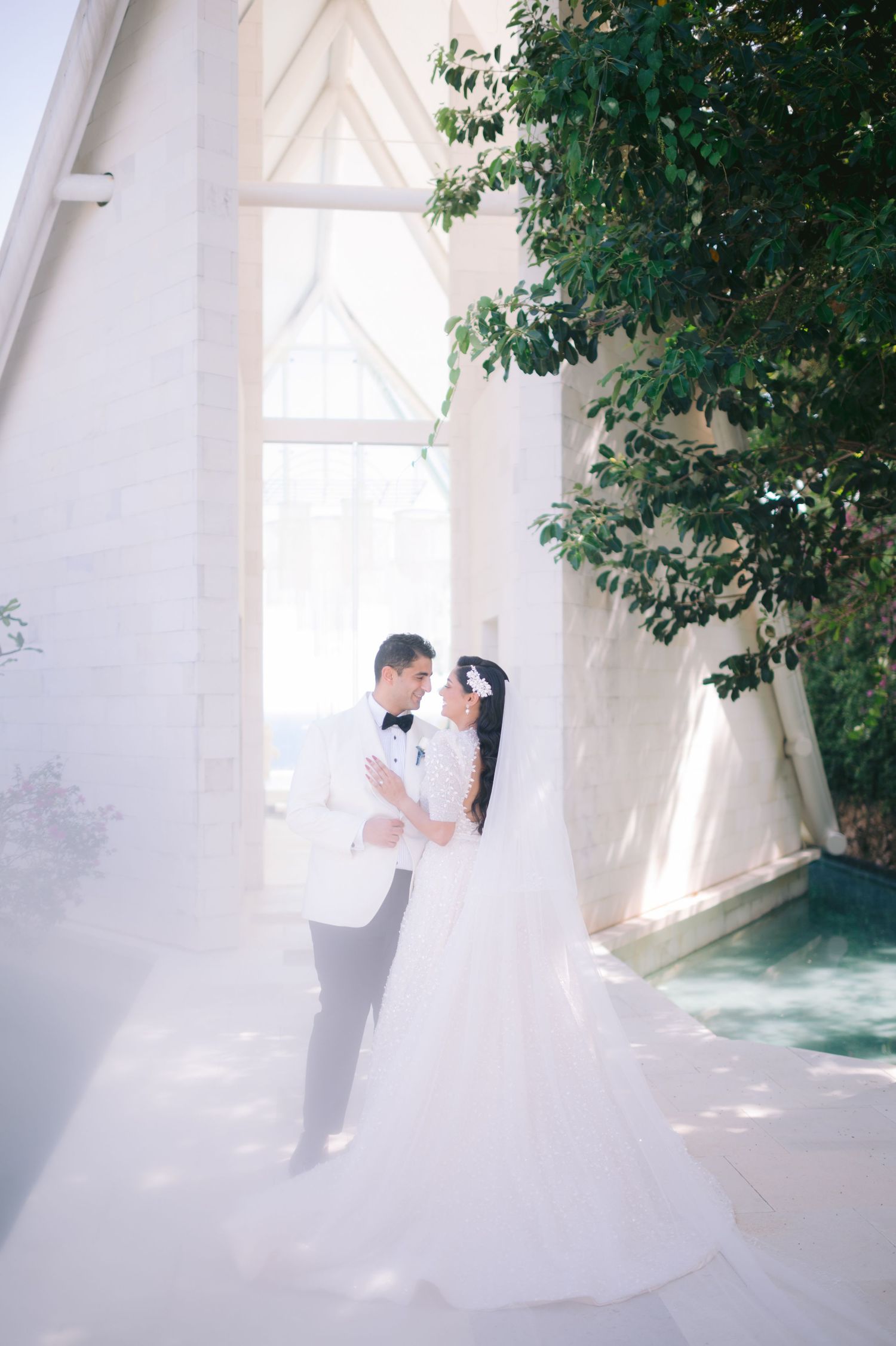 A bride and groom share a romantic embrace outside a white chapel, her veil flowing gracefully in soft natural light.