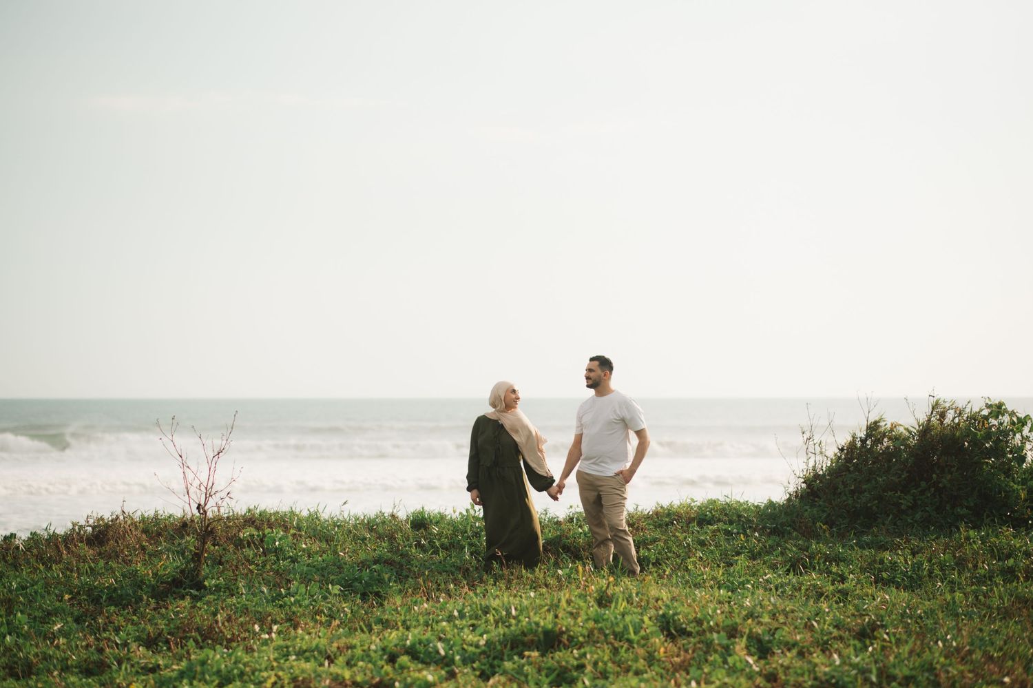 A couple holds hands on a lush green coastal cliff overlooking ocean waves on a bright day.
