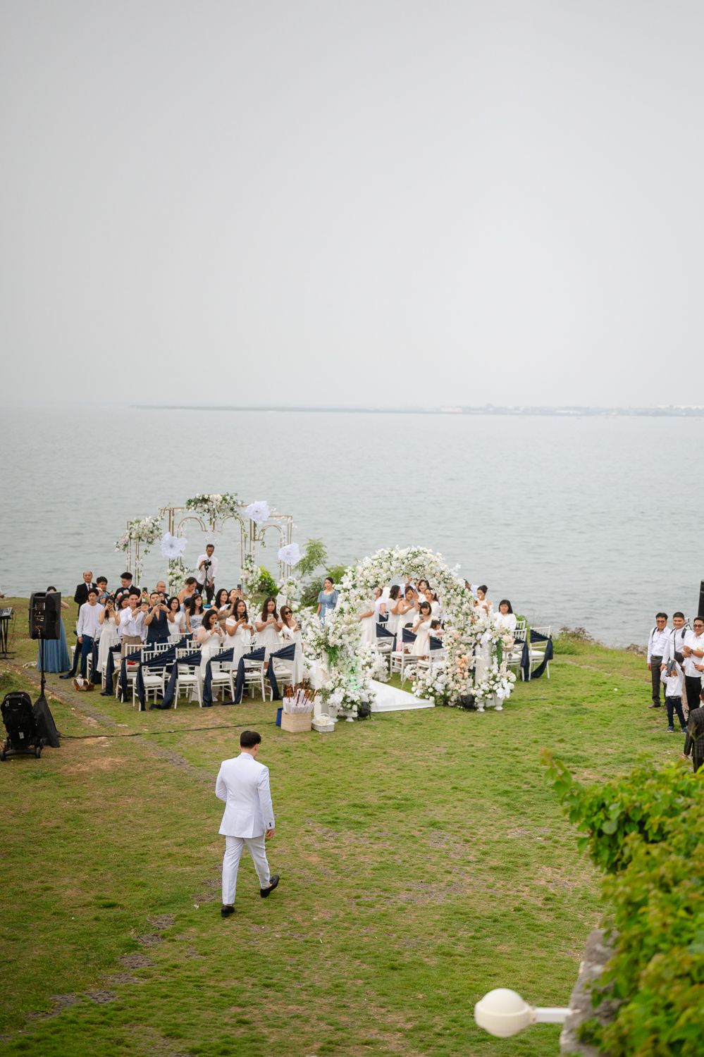 Elegant outdoor wedding ceremony by the sea with white floral arch, guests in white attire seated on green lawn.