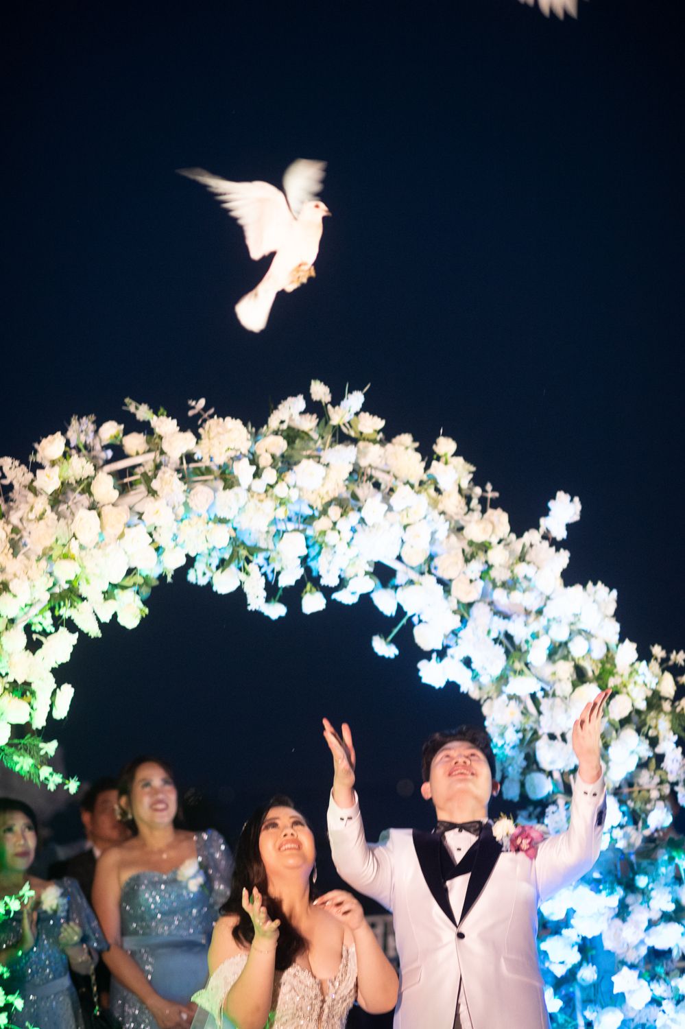 Newlyweds release white doves under a floral arch at night, with guests watching in celebration.
