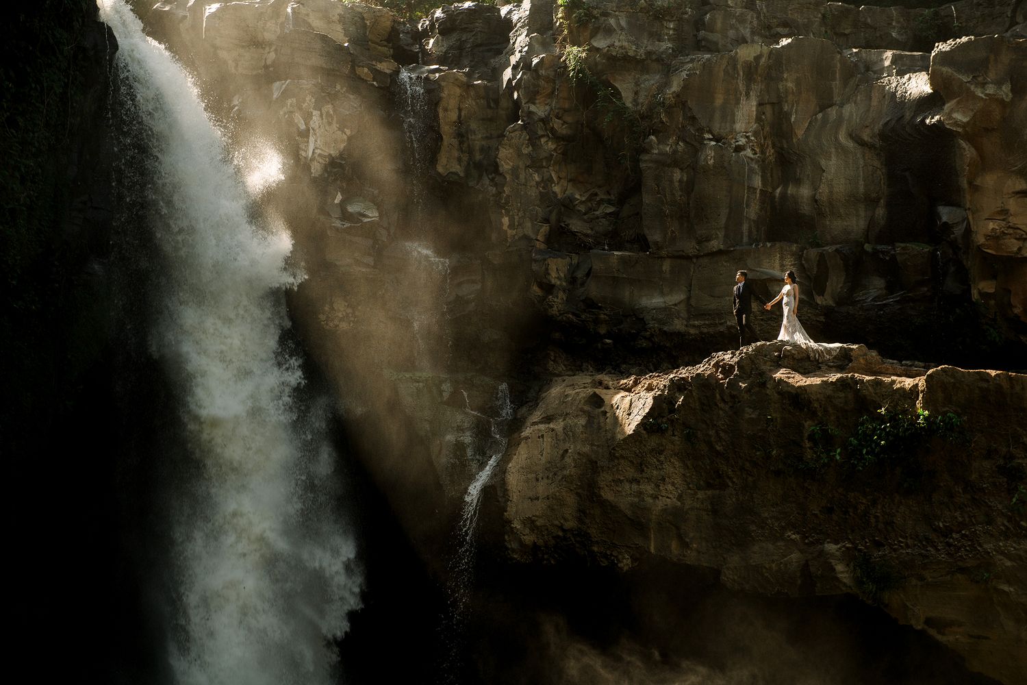 Couple standing on rocky ledge beside a dramatic misty waterfall in a dark canyon.