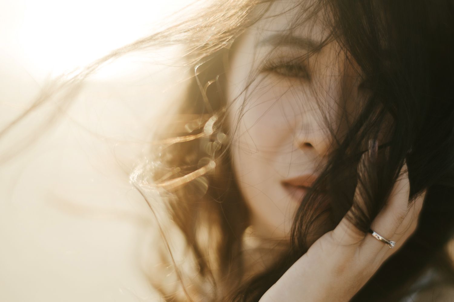 A woman with flowing dark hair caught in sunlight, eyes closed, wearing a silver ring, in a dreamy artistic portrait.