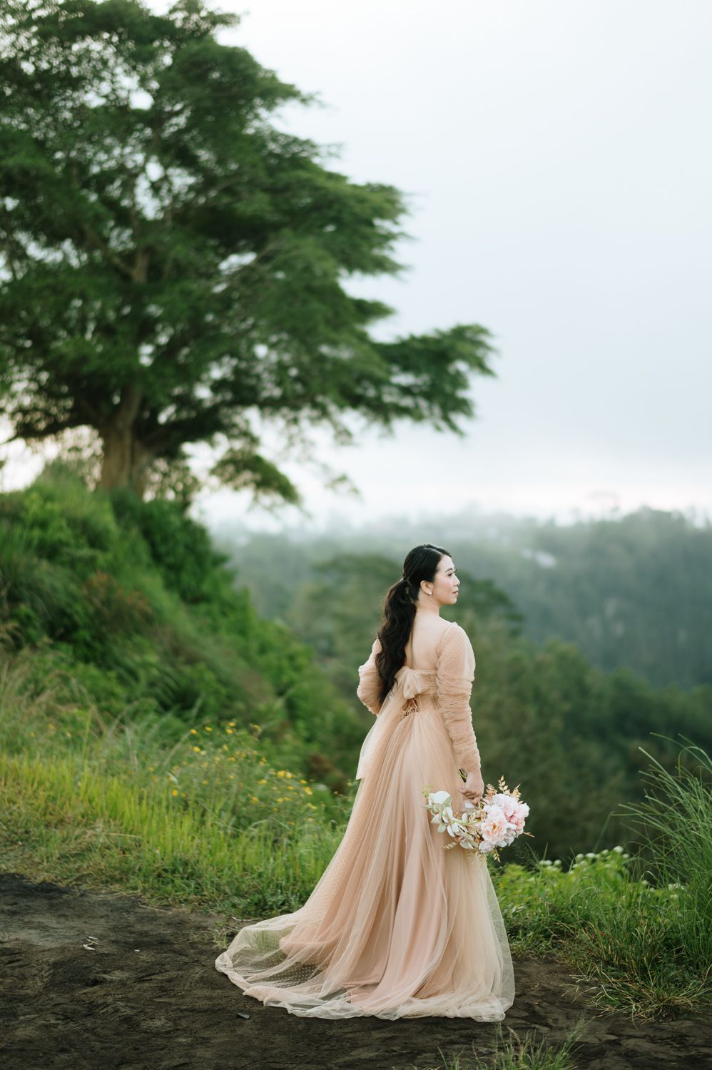 A bride in a flowing champagne tulle gown holds a bouquet while standing on a misty hillside with lush greenery.