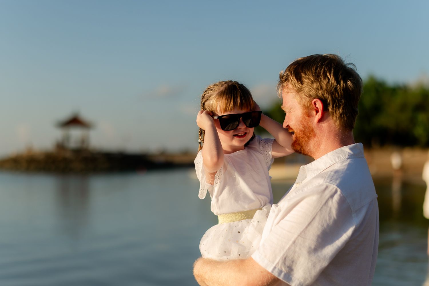 Father holding young child wearing oversized sunglasses on a sunny tropical beach at sunset.