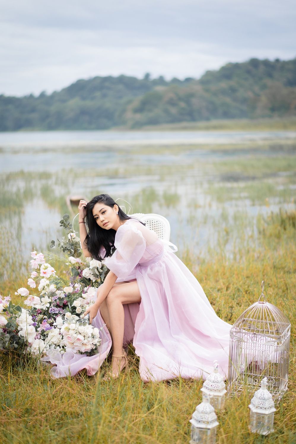 Woman in pink gown poses gracefully in a meadow near a lake with flowers and a decorative birdcage.