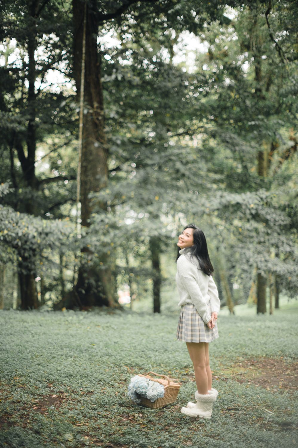 Young Asian woman in plaid skirt and white sweater stands smiling in a lush green forest with a wicker basket nearby.