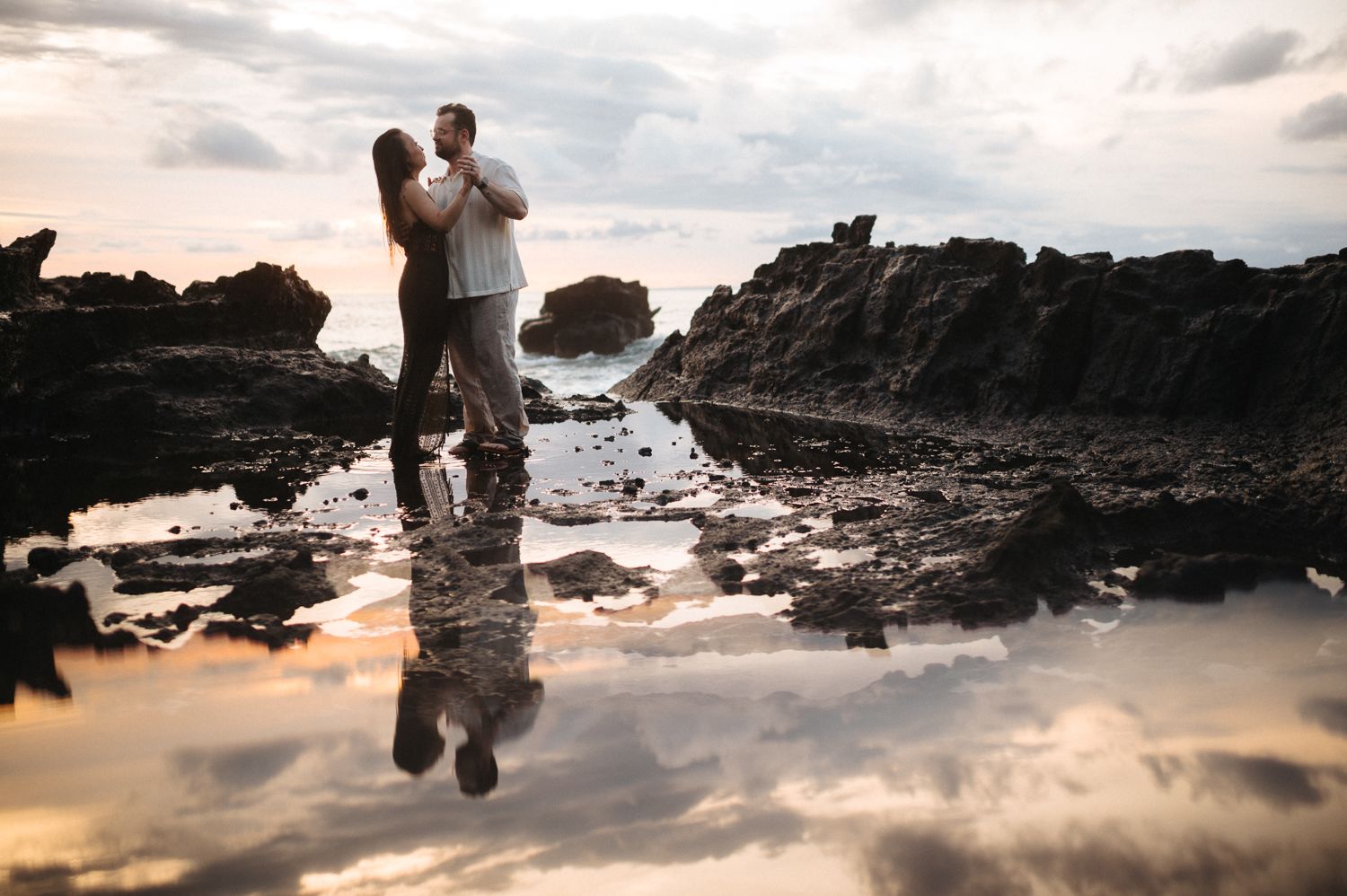 Couple embracing on rocky coastal shore at sunset with stunning reflection in tide pool water.