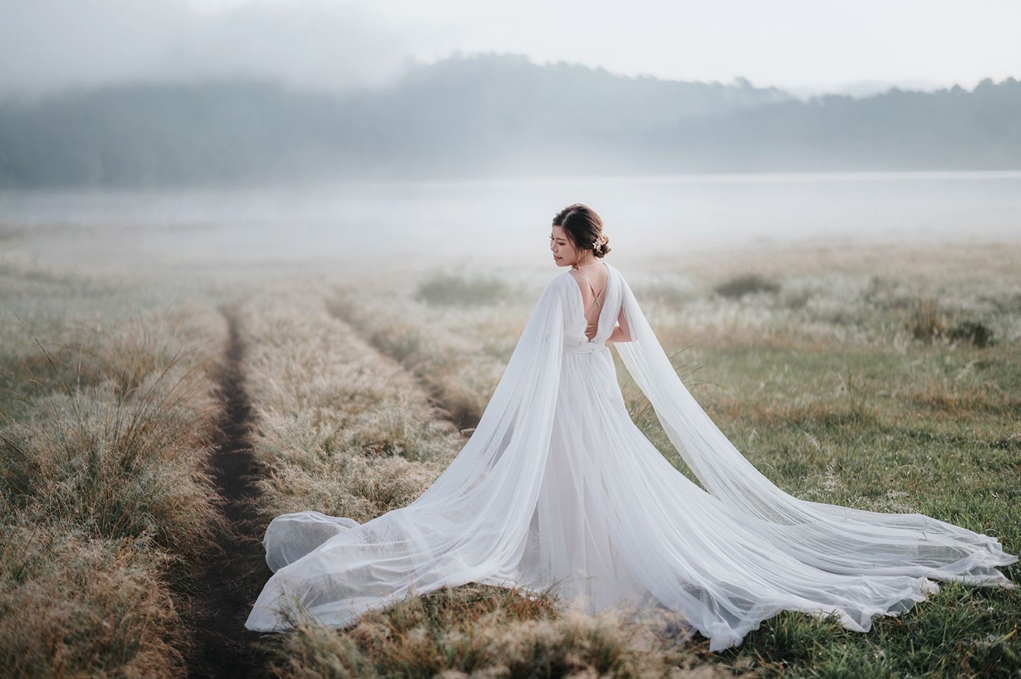 Bride in flowing white gown with cape train stands in misty field at dawn.
