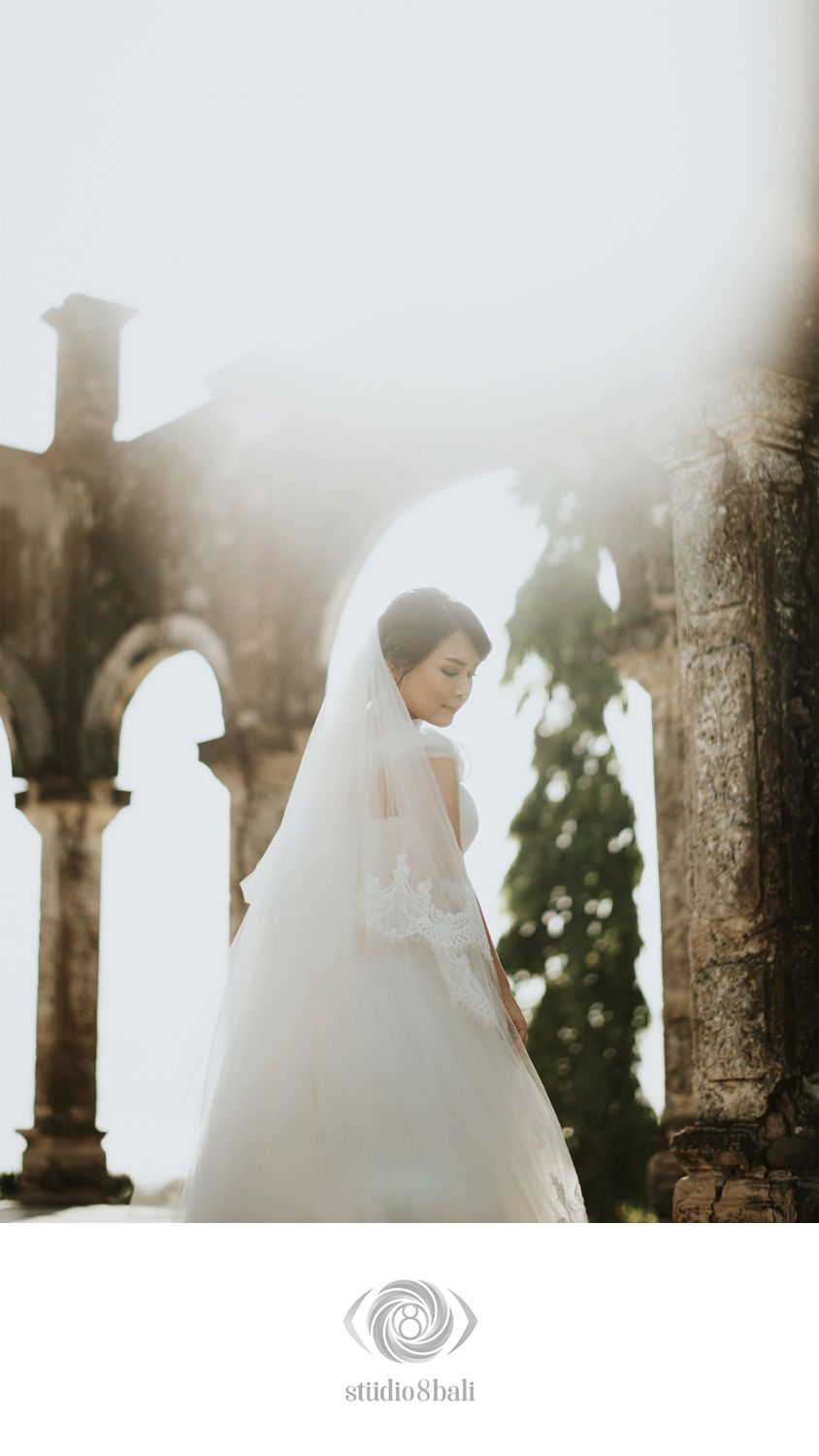 Bride in white gown and veil stands gracefully before ancient stone arches in soft golden light.