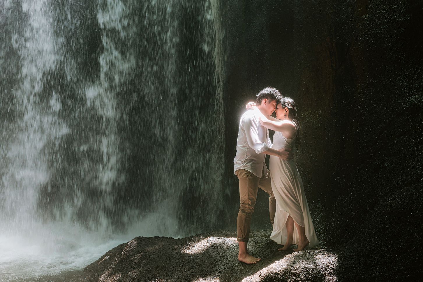 Romantic couple embracing in front of a stunning waterfall, standing on rocks in elegant white attire.