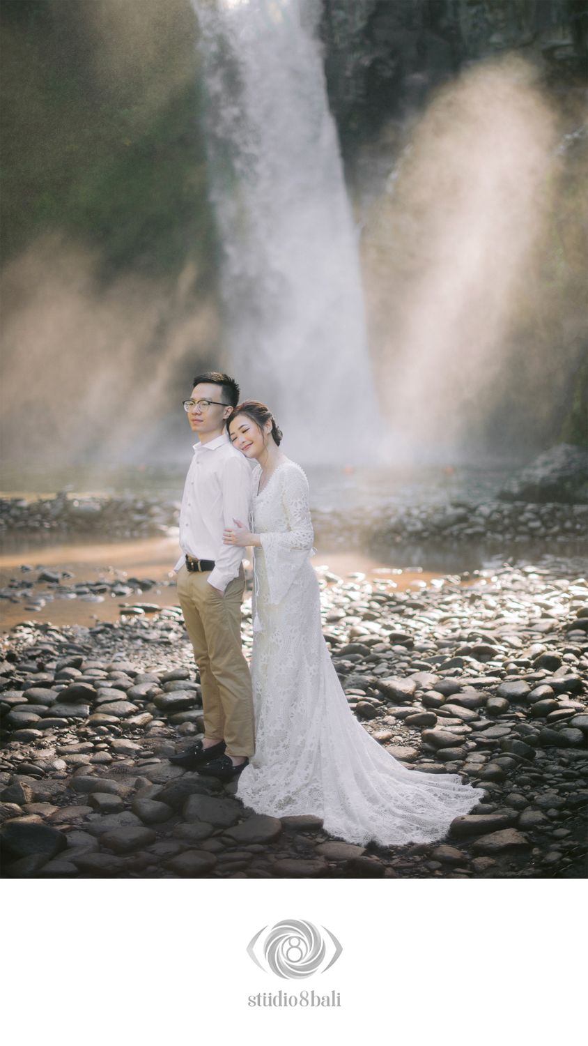 Couple in wedding attire poses romantically on rocky riverbank before a majestic waterfall in Bali.