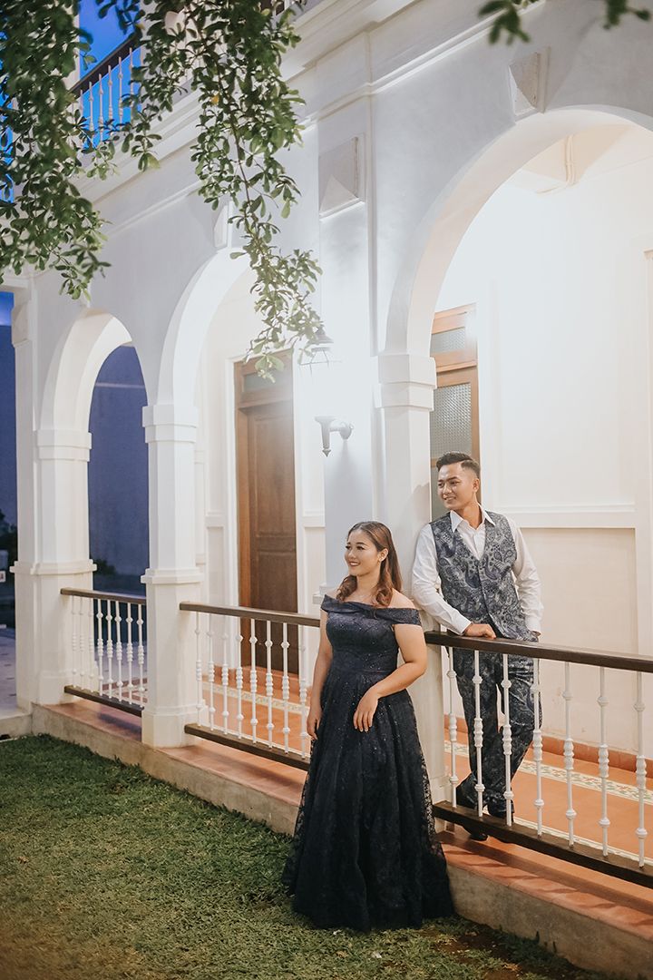 Couple in elegant formal wear posing on a white colonial-style building veranda at dusk.