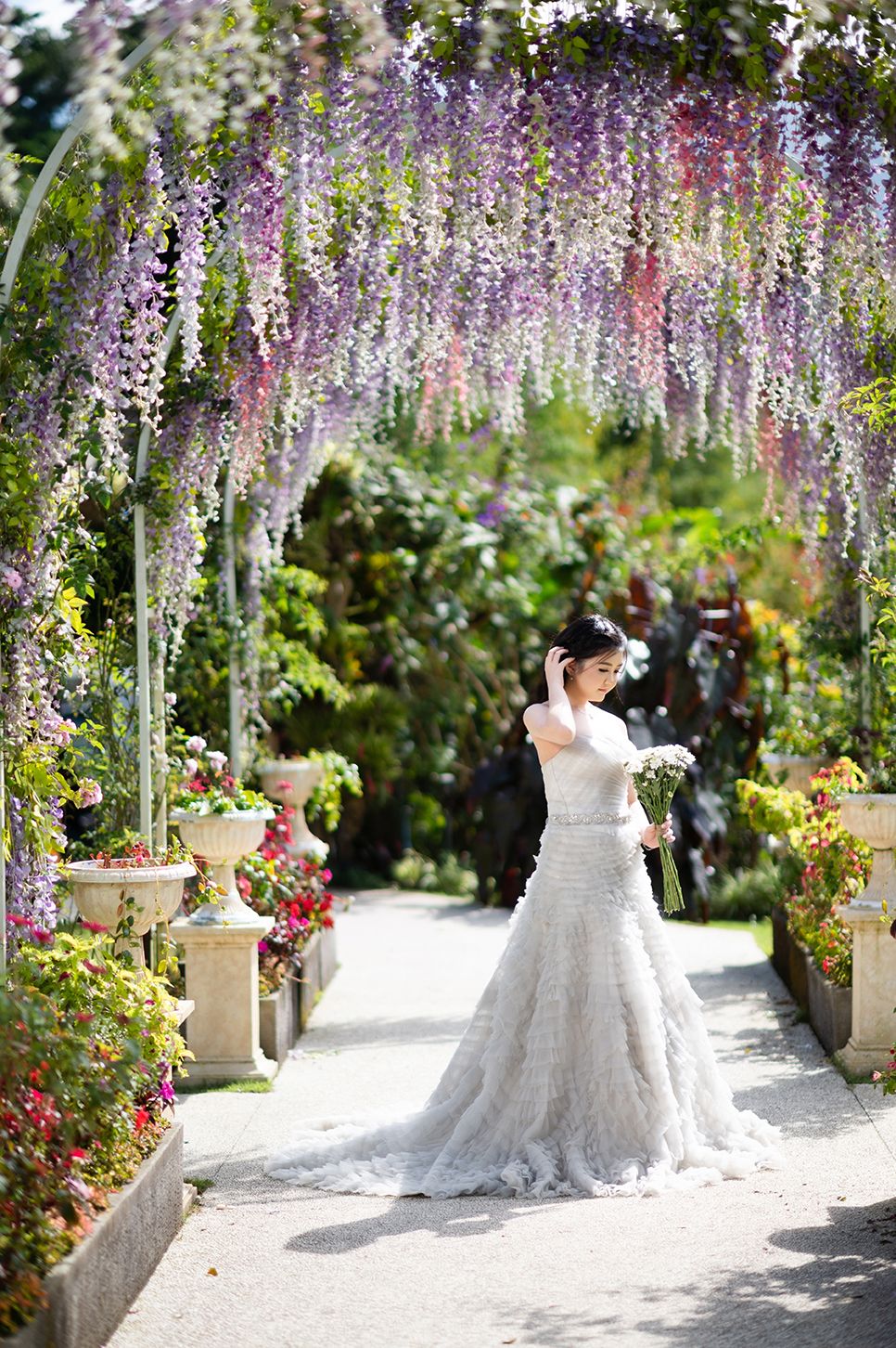 Bride and groom stand under a stunning wisteria archway in a lush garden, bride wearing an elegant lace mermaid gown.