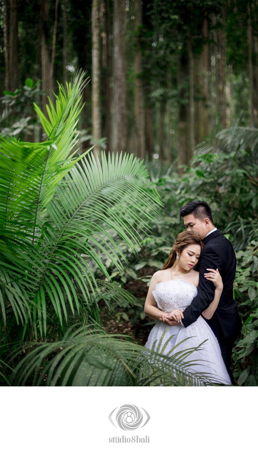 Couple in wedding attire embrace romantically among lush tropical palm leaves in forest.