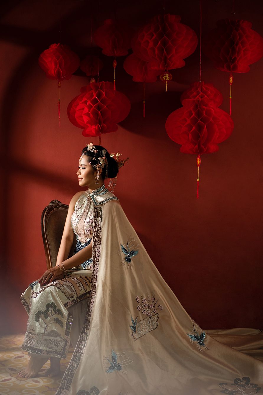Woman in elegant traditional Chinese attire with embroidered cape sits gracefully beneath red paper lanterns.