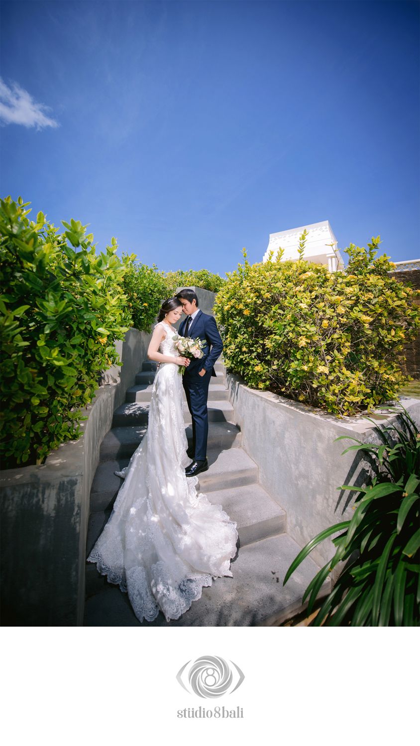 Bride and groom share a romantic kiss on garden stairs, bride in lace gown holding bouquet under a bright blue sky.