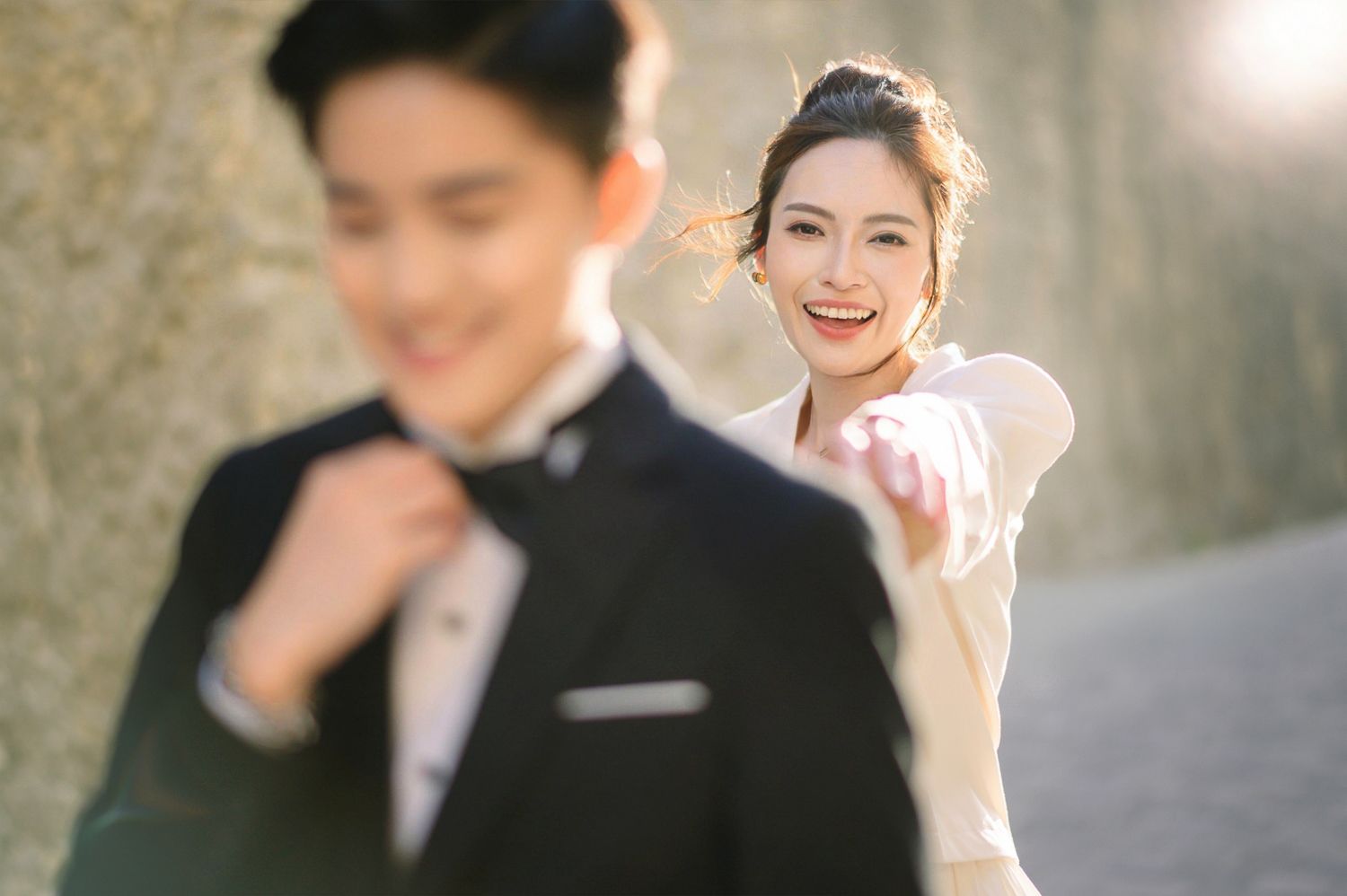 A smiling bride reaches toward the camera as her groom adjusts his suit in the foreground.